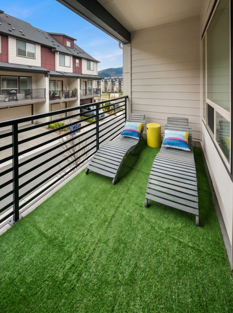 Balcon mis en scène : gazon synthétique, deux transats en bois, table d'appoint jaune, coussins colorés et vue sur les façades.