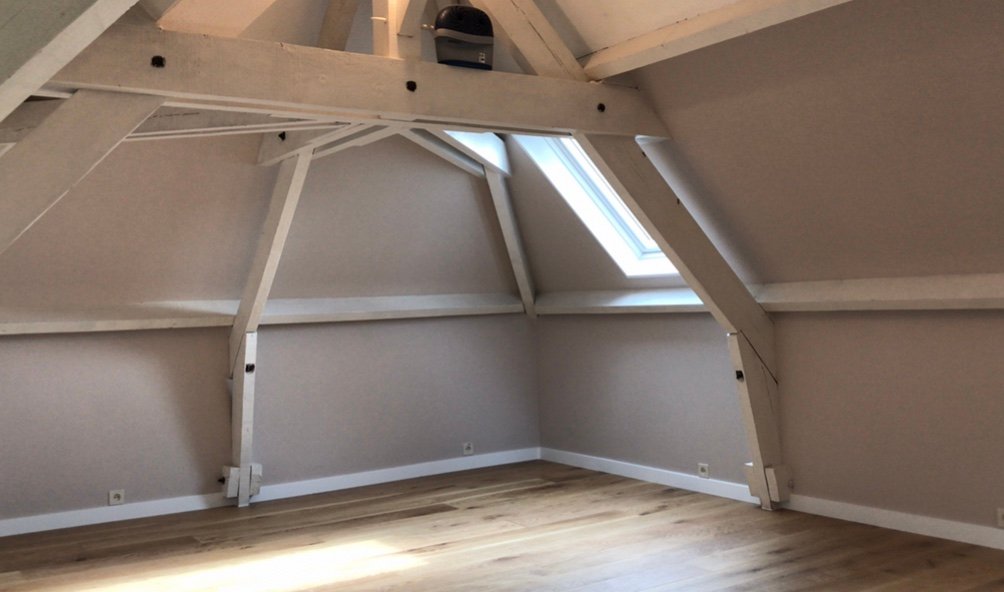 Combles avant aménagement : charpente apparente, murs clairs, sol en bois et velux, pièce vide.