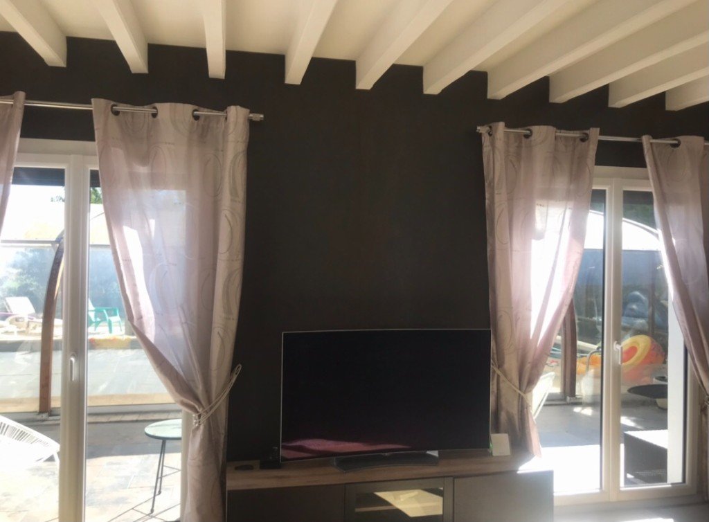 Mur TV avant réaménagement : fond sombre, meuble bas et baies vitrées avec rideaux clairs.